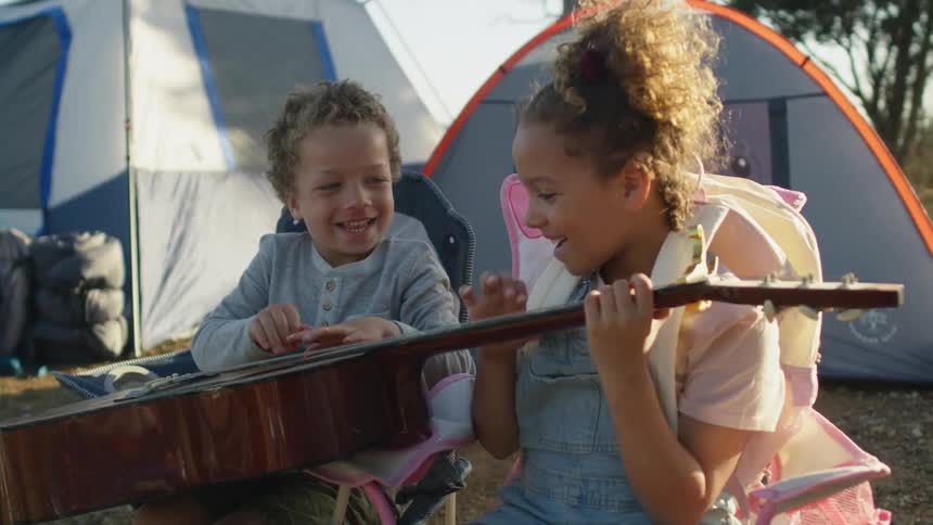 A young brother and sister attempt to play a guitar together on a campsite during the day