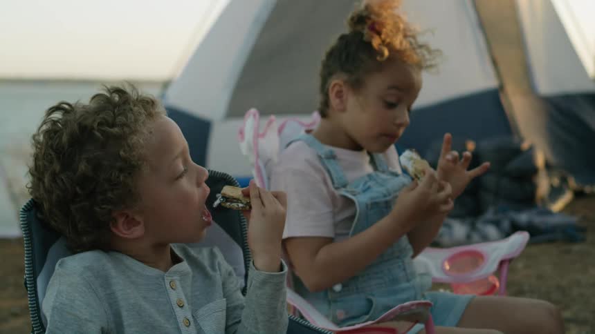 Two children are sitting in a campsite and eating smores