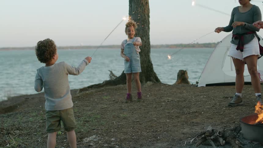 A family of four standing at their campsite waving sparklers near the lake