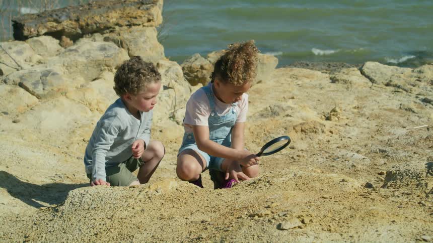 A little boy watches closely as his sister uses a magnifying glass to look at rocks on shore