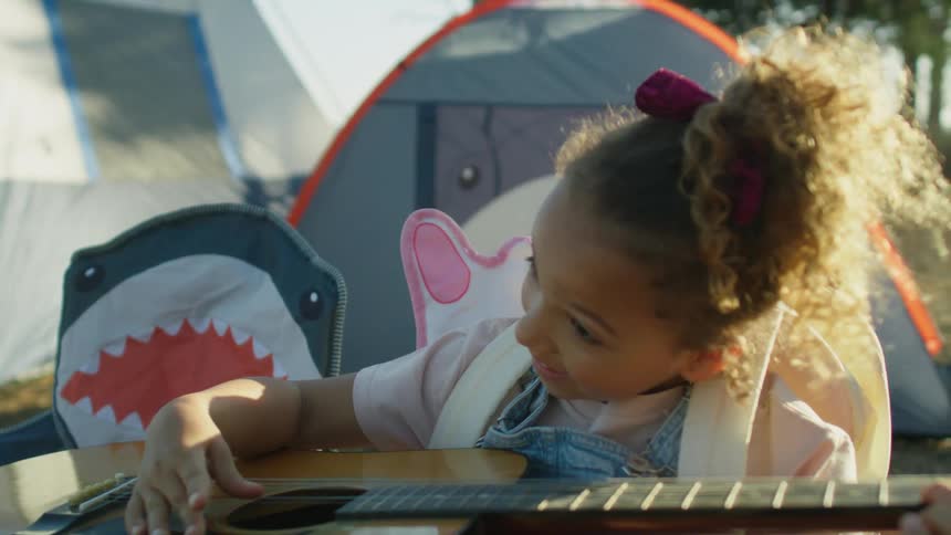 A little girl smiles as she sits in a chair near her tent pretending to play a guitar