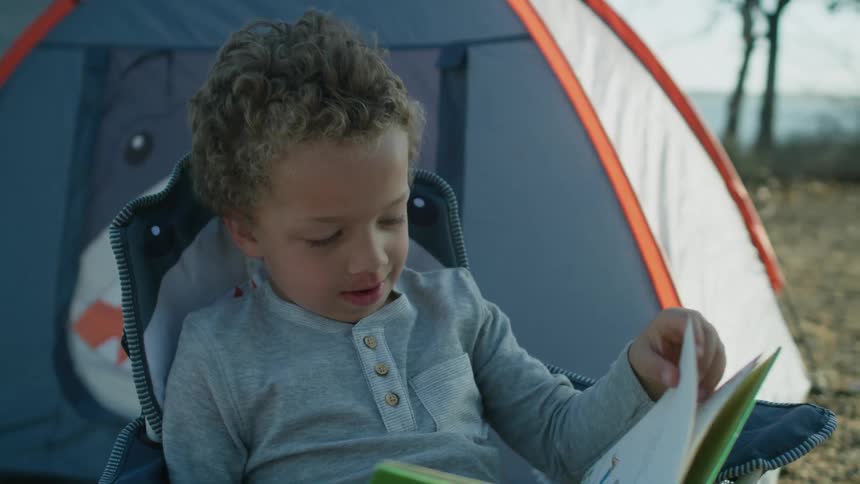 A little boy is sitting outside of a tent looking at a book