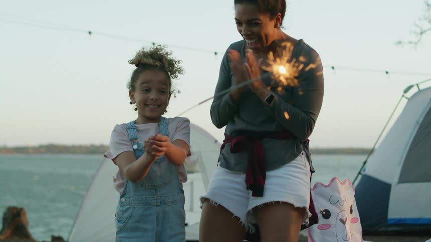 A woman cheers while her daughter waves a sparkler around at their campsite by a lake