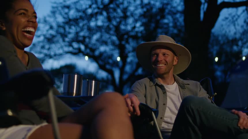 A man and woman laugh as they chat with one another while sitting outdoors on a campsite in the evening