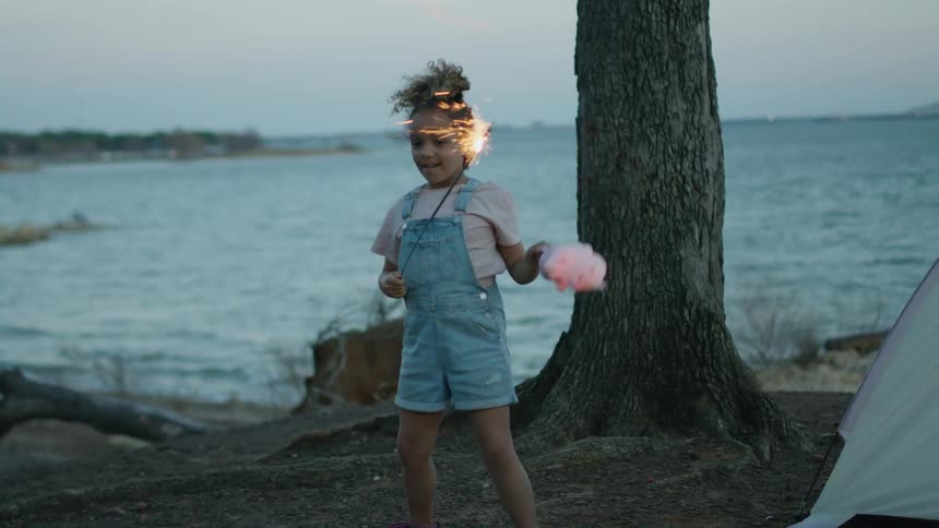 A young girl dances next to her tent with a sparkler in one hand and cotton candy in the other