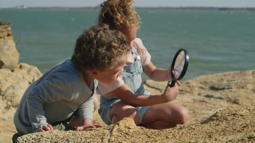 Siblings look through a magnifying glass while sitting along the shore of a lake