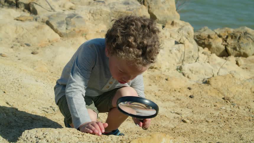 Child crouches on beach examining ground with a large magnifying glass