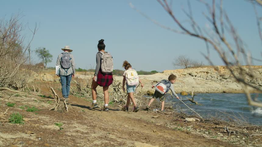 A boy plays with a stick in the water as his family hikes past a lake