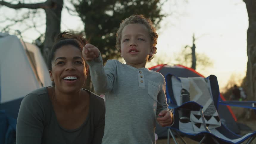 Mom smiles and encourages her little boy as he holds and swings a sparkler at the campsite