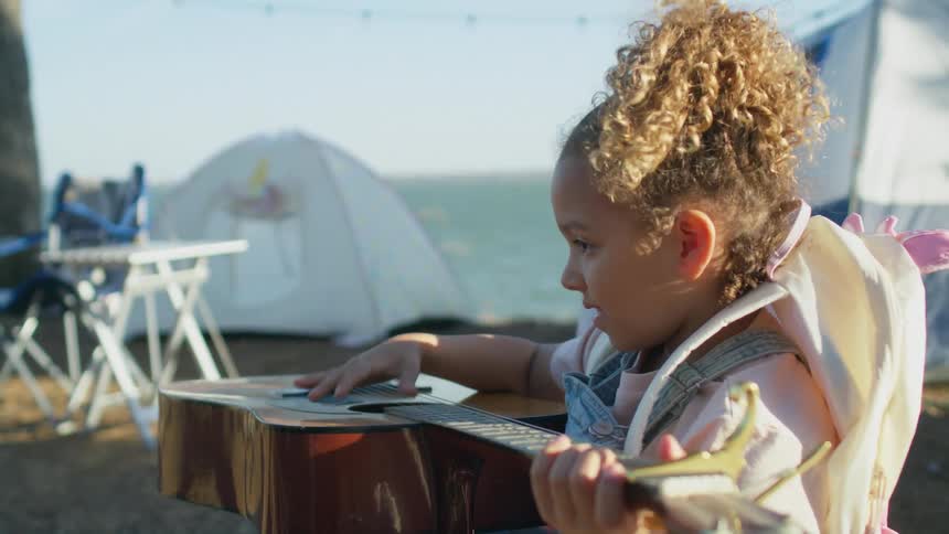 A little girl sitting in a camp chair strumming on a big guitar with tents and lake behind her