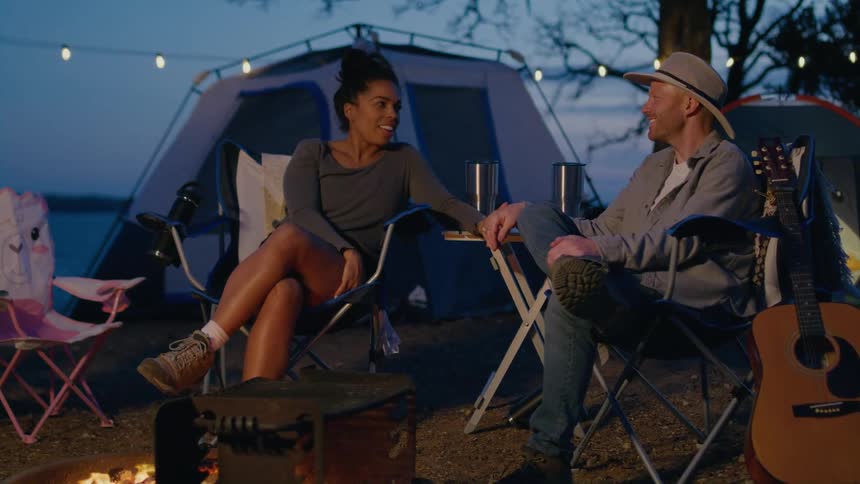 A man and a woman are sitting outside near a campfire and a tent along the coast holding hands and he has a guitar next to him