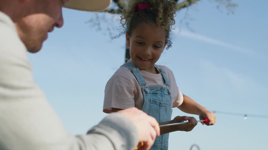 A young girl camping with her dad takes a hot dog off a grill and puts it in a bun he is holding open