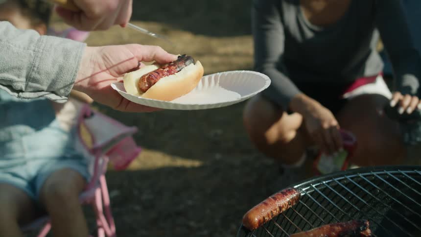 A man camping grills hotdogs and puts one on a bun and passes it to his wife