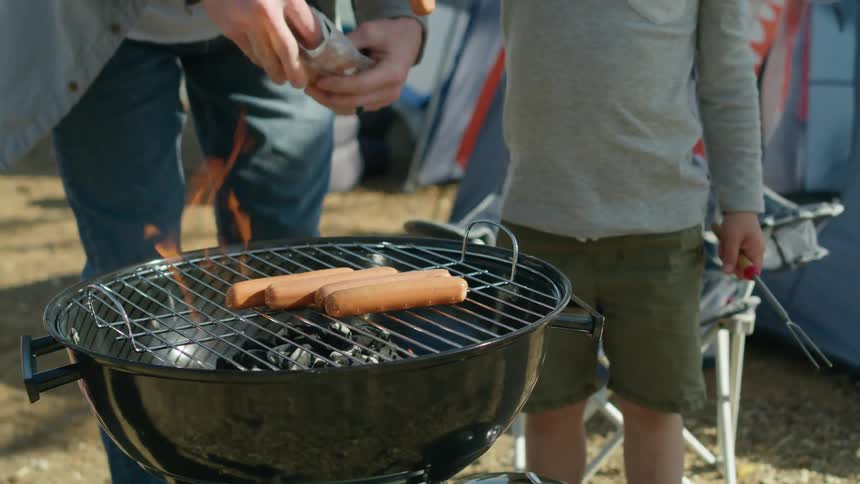 A man camping with his son puts hotdogs on a grill