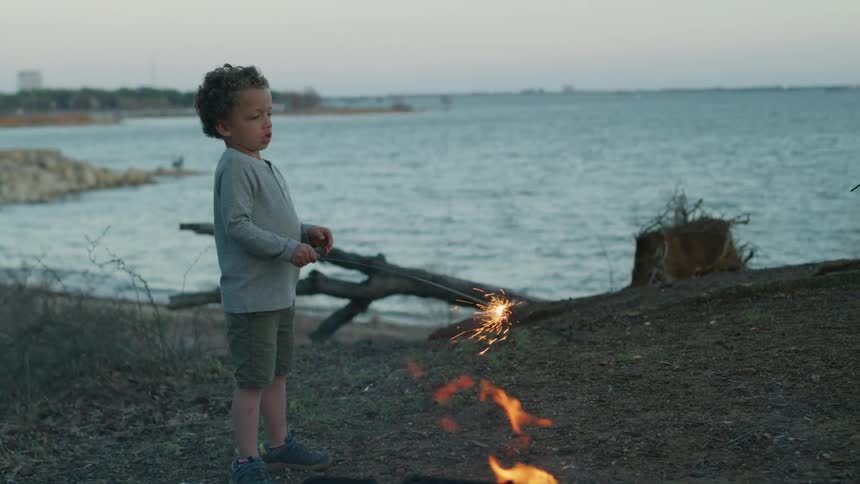 A young boy leisurely twirls a sparkler while standing on the shore of a lake in the evening