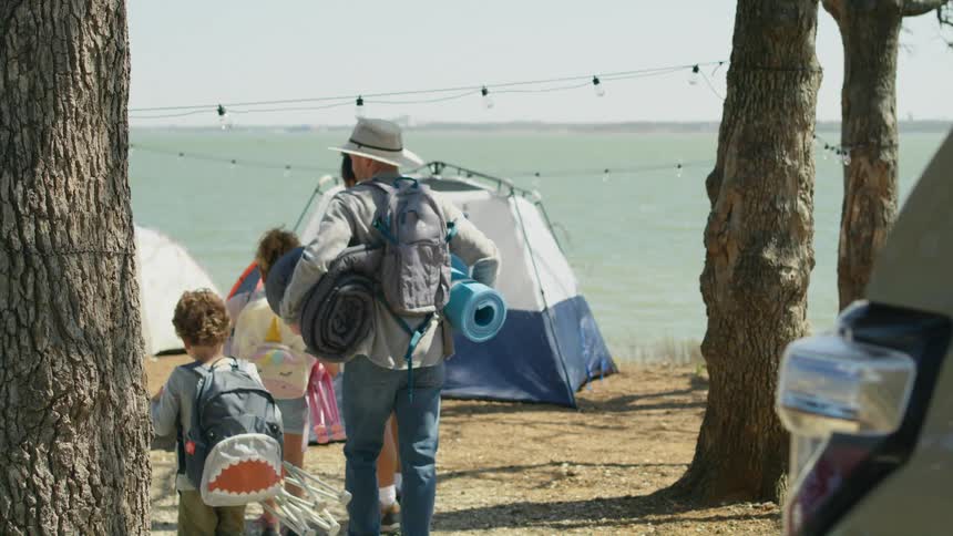 A family carries equipment from a car to tents on a campsite