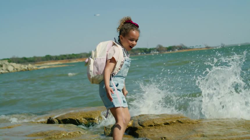 A little girl wearing a backpack is standing near the water and when it splashes on her she turns and looks at her little brother