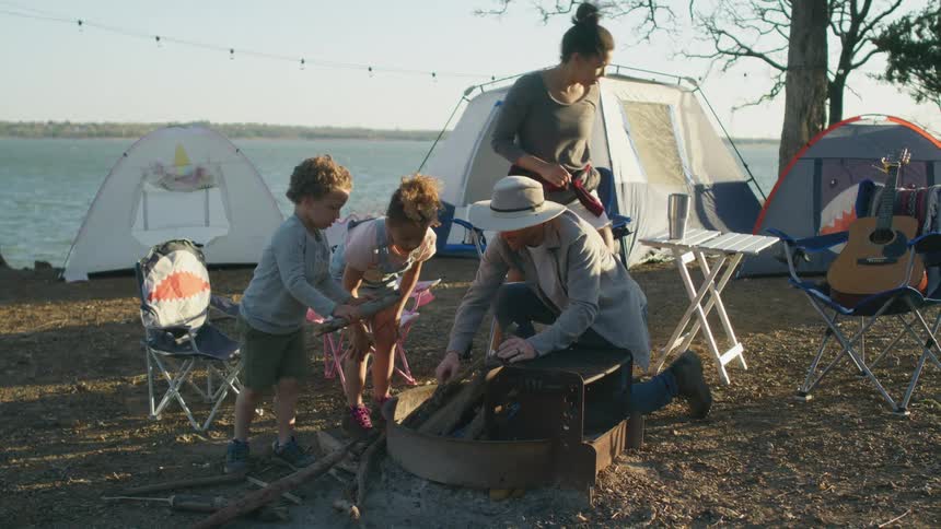 A little boy and girl put sticks of wood in a fire pit as they help their dad build a fire while camping