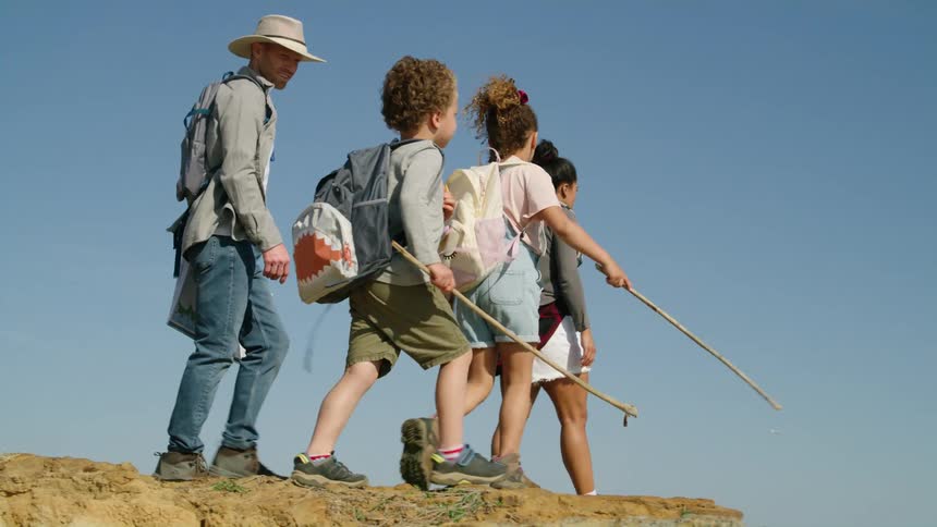 A family with two young children all wearing backpacks are hiking across a mountain ridge under a clear blue sky
