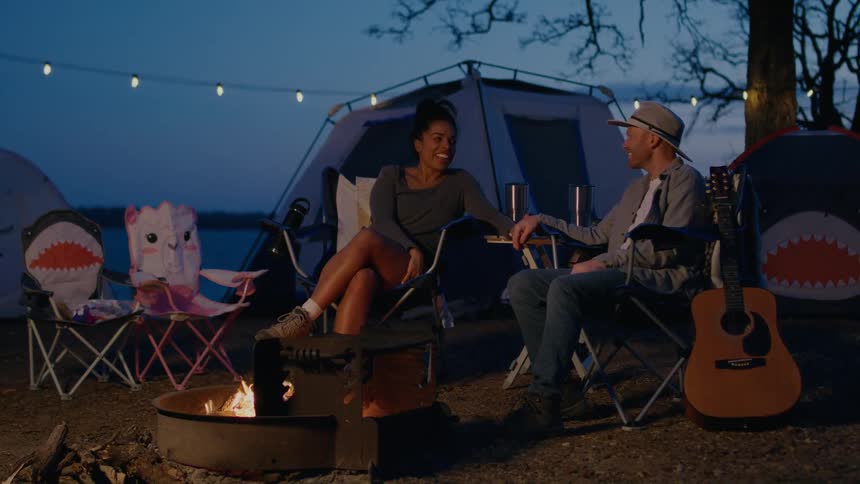 A man and woman chat with one another while sitting beside a campfire at a campsite on a lakeshore at night