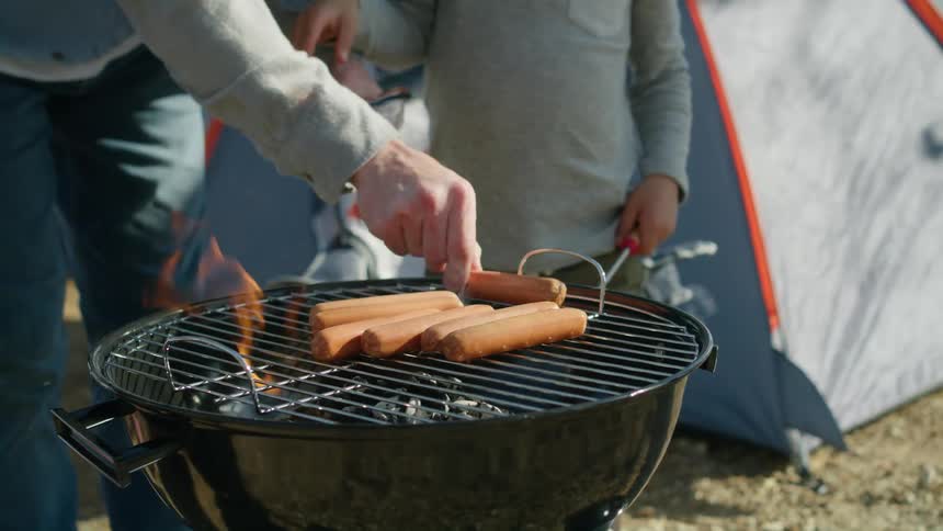 Father and son grilling sausage on the open flame of a barbecue while camping