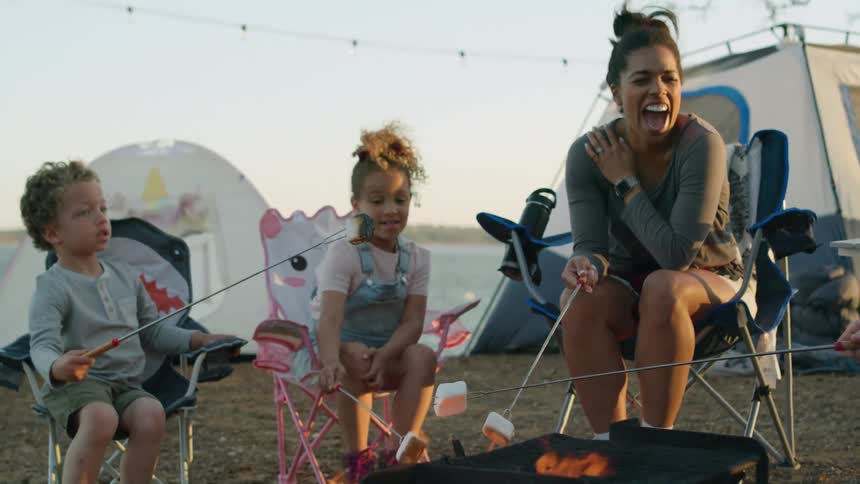 A family cooks marshmallows over the flames of a grill on a lakeshore campsite during the day