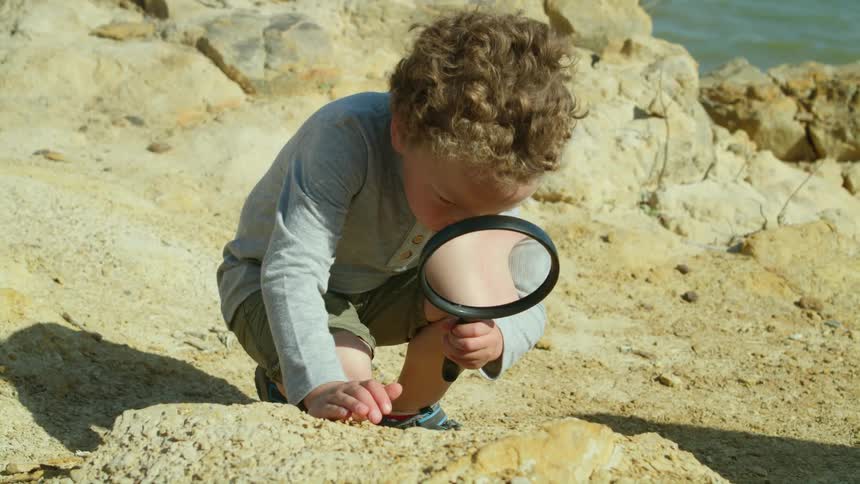Child crouches on shore near water to examine ground with a magnifying glass