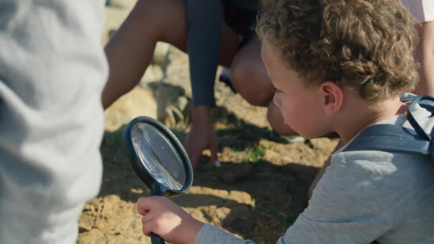 A little boy is outside with other people and he is holding a magnifying glass over the ground