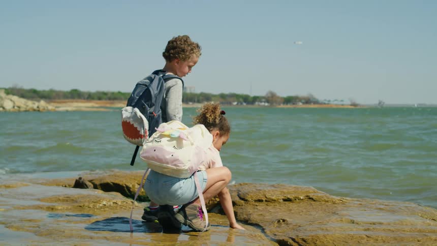 A little girl kneel on the shore and puts her hands in the water as her brother walks towards the water