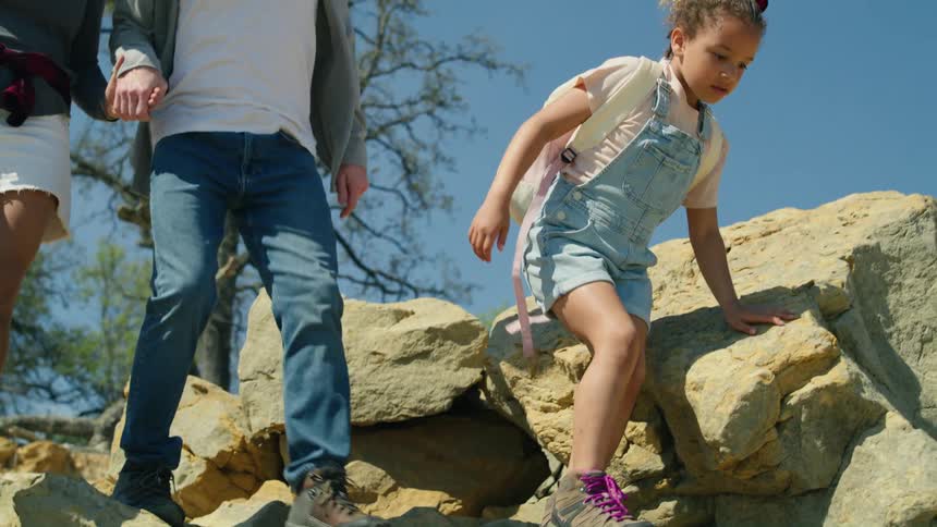 A little girl is walking on rocks along the side of a hill when she jumps down and her parents are walking behind her