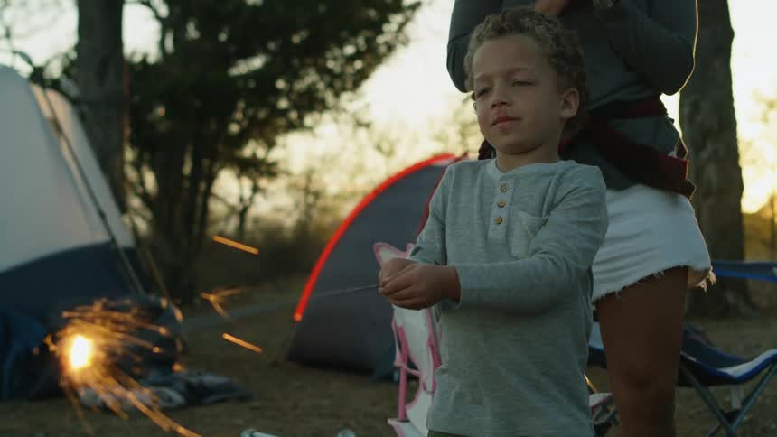 A little boy is standing outside near tents holding a sparkler and his mother is behind him