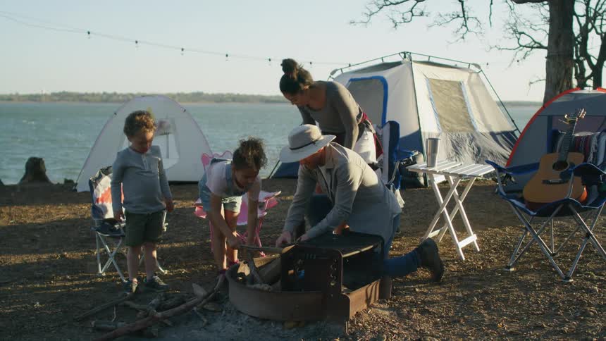 A little girl adds a stick to the fire ring helping dad at their campsite