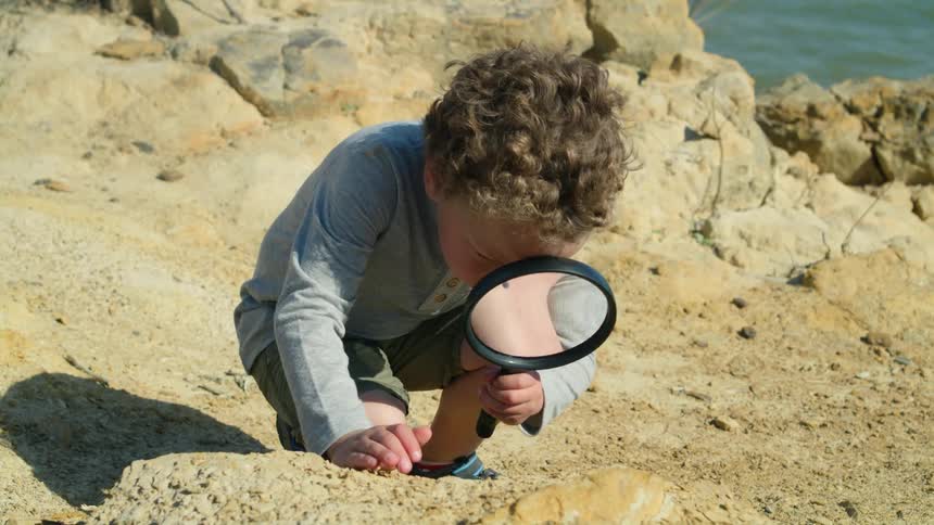 A young boy on the shore holds a magnifying glass and looks at the rock