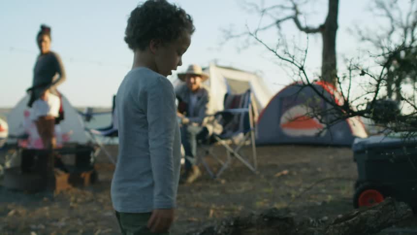 A young boy camping with his family carries a big piece of wood and drops it in a pile as his father who is playing a guitar and his mother watch