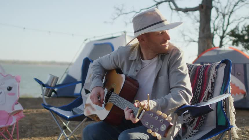 A man camping by a lake sits in front of tents and plays the guitar