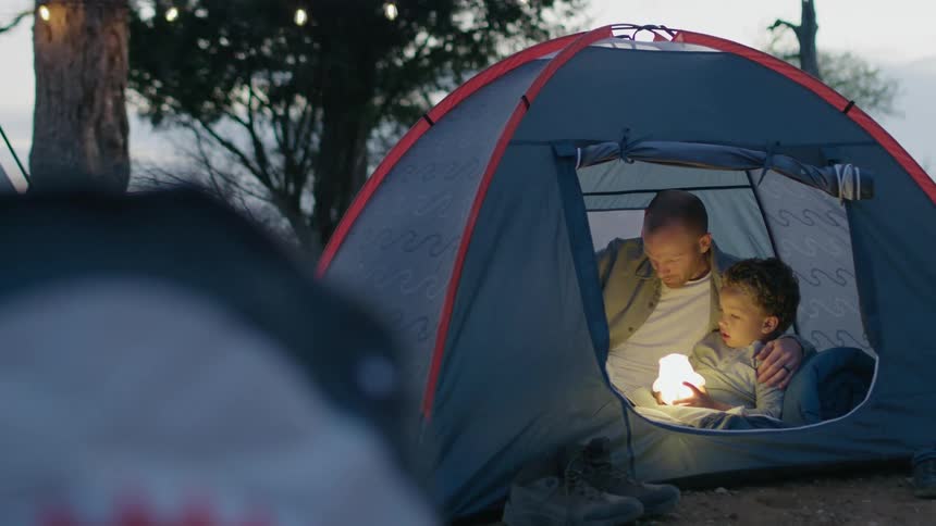 Father and son sit in tent with a camp light on a windy day reading