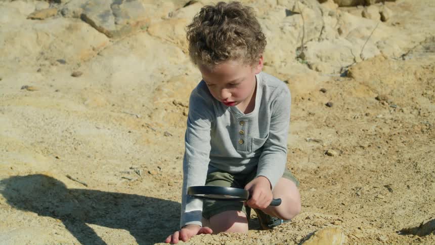 A young boy kneeling outdoors uses a magnifying glass to examine the soil during the day