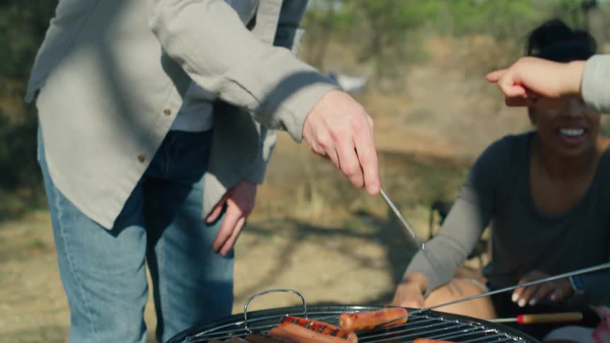 A man grills hotdogs outside while others hold sticks to grill with