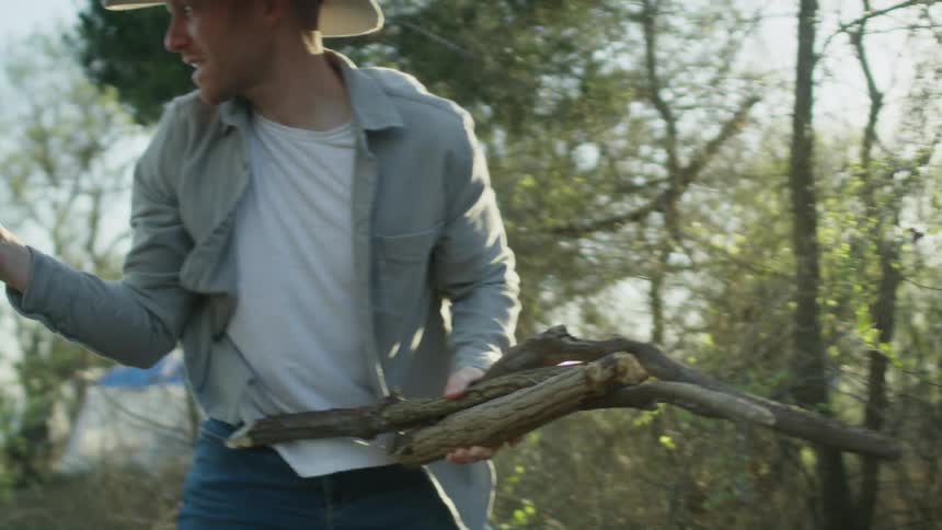 A man out camping picks up pieces of wood in a forest and hands them to somebody else