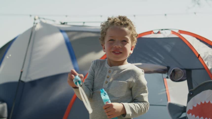 A boy blows bubbles in front of tents outside