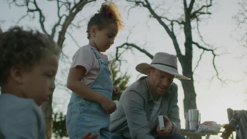 A man camping with his son and daughter shows them a long match and hands it to his daughter