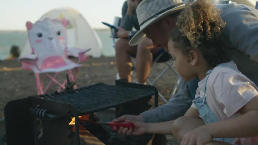 A dad is helping his daughter with a stick in the fire at a campsite while the mom looks on