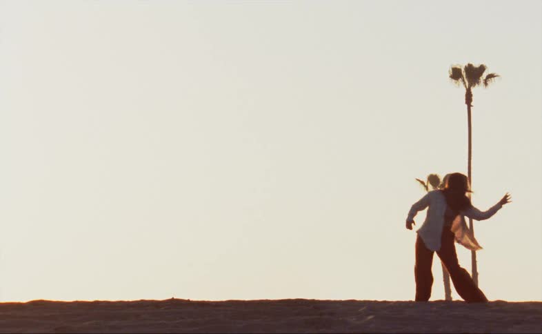 A woman wearing red baggy pants and a white shirt is outside dancing near palm trees