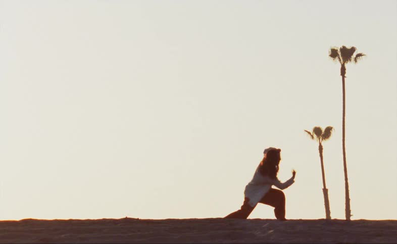 A woman wearing red baggy pants and a white shirt is outside dancing near palm trees