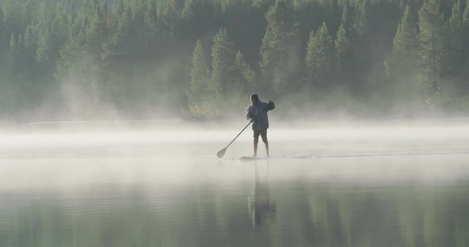 A man is on a board rowing across a fog covered lake