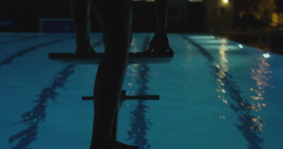 Woman climbs on top of a diving platform overlooking a swimming pool at night