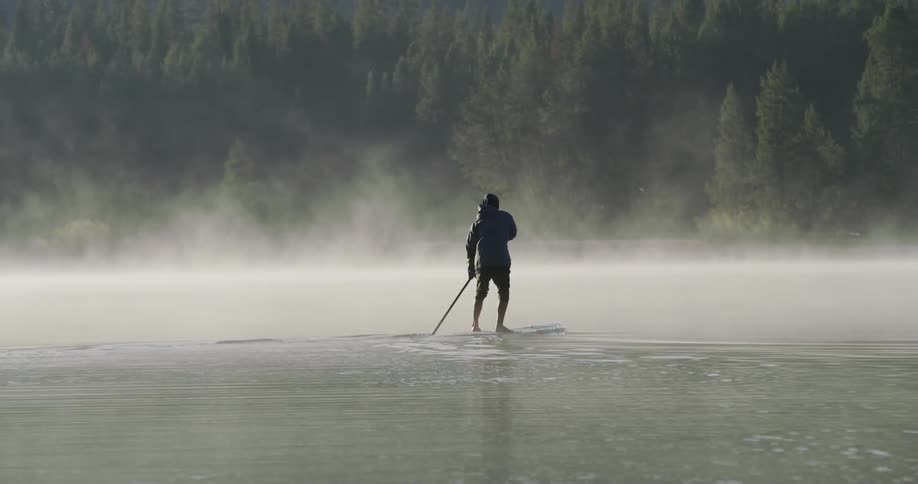 A man paddles a paddle board over a mist covered waterway