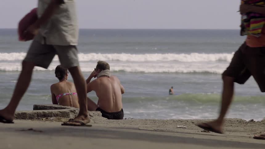 A couple sit on the beach while others walk behind them