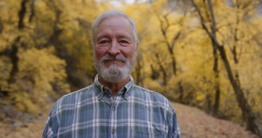 An elderly man smiles into the camera while standing on pathway through a woodland on an autumn day