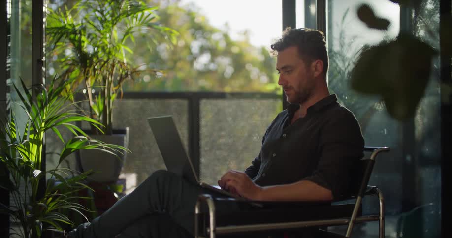 Man with beard and navy blue shirt sits armchair uses laptop in living room